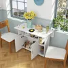 A white folding table with cabinets is set up next to a window, adorned with a vase of yellow flowers, a bowl of snacks, and a bottle of oil. Two wooden chairs are placed around the table. Voir les images du produit