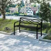 A black metal park bench sits on a paved patio in front of a body of water with houses in the background. Produktbild-Vorschau