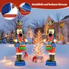 Two illuminated nutcracker figures with drums stand in a snowy yard in front of a house, with a small decorated tree between them. Podgląd zdjęcia produktu