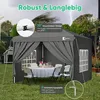 A grey gazebo set up in a garden with a table and chairs inside, under a stormy sky. Produktbild-Vorschau