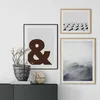 Three framed pictures are displayed on a white wall above a dark cabinet. A plant in a basket sits on the cabinet to the left, next to a wooden decorative object. Podgląd zdjęcia produktu