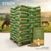 A large stack of feed bags on a pallet, with a single bag and spilled feed in the foreground, and horses in a field in the background. Produktbild-Vorschau