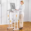 A woman is standing at a white adjustable height desk, working on a computer. The desk has wheels and a small drawer. Orange arrows indicate the desk's adjustable height. Podgląd zdjęcia produktu