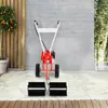 A red and white tiller/sweeper machine is positioned on a patterned rug in front of a brick wall and a wooden fence. Produktbild-Vorschau
