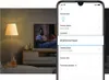 A smartphone screen displaying a smart bulb's settings menu, with a lamp and TV in the background. Podgląd zdjęcia produktu