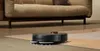 A robot vacuum cleaner is cleaning a rug in a living room with a sofa in the background. Voir les images du produit