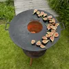 A round grill plate with sliced figs arranged on it, placed on top of a fire pit in a grassy outdoor setting. Produktbild-Vorschau