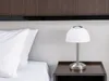 A close-up of a bedside table with a lamp and pillows on a bed. Produktbild-Vorschau