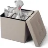 A beige storage ottoman with its lid open, revealing a teddy bear and two plaid pillows inside. The ottoman is placed on a white background. Produktbild-Vorschau
