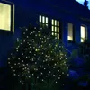 A bush decorated with warm white LED lights at night, in front of a dark house with illuminated windows. Produktbild-Vorschau