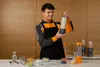 A man in an apron is holding a blender bottle, with other blenders and ingredients on a counter. Produktbild-Vorschau