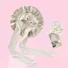 A beige silk bonnet with long ties is displayed against a pink background, with a small dish of silkworm cocoons and a decorative postcard nearby. Voir les images du produit