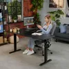 A woman is working at a black L-shaped standing desk in a home office setting. Produktbild-Vorschau