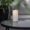 A white LED candle sits on a glass-topped table outdoors, with a blurred green plant and furniture in the background. Produktbild-Vorschau