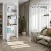 A white bookshelf filled with decorative items and books, placed in a living room setting next to a sofa and a large plant. Náhled obrázku produktu