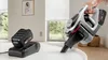 A hand holds a cordless vacuum cleaner near a table, with a charging battery in the foreground. Náhľad obrázku produktu