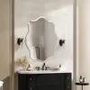 A modern bathroom vanity with a black cabinet, white countertop, and a uniquely shaped mirror above it. Two wall sconces flank the mirror, and decorative items are placed on the counter. Anteprima dell’immagine prodotto