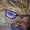 A close-up of a watercolor painting of an eye, with a paintbrush adding detail to the iris. The painting features purple and white floral elements. Voir les images du produit