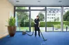 A woman is vacuuming a blue carpet in a modern office space with large windows overlooking a garden. Produktbild-Vorschau