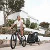 A man rides a bicycle with a trailer attached, in a residential area with modern architecture. Produktbild-Vorschau