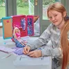 A young girl is shown at a table with a Barbie fashion salon playset and doll, surrounded by fabric and sewing patterns. Produktbild-Vorschau