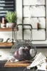 A wire basket filled with red and white onions sits on a wooden cutting board, with a striped cloth to the side. The background shows shelves with plants and kitchen items. Produktbild-Vorschau