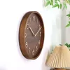 A wooden wall clock with gold numbers and hands is shown mounted on a white wall, with a pleated lampshade and green leaves visible in the background. Voir les images du produit