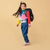 A young girl wearing a colorful sweater, jeans, and a backpack, posing against a plain background. Anteprima dell’immagine prodotto