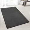A dark gray rug is laid out on a light wooden floor in a well-lit room with sheer white curtains in the background.