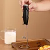 A hand holding a black electric milk frother over a glass mug of milk, with a wooden tray and coffee beans in the background. Produktbild-Vorschau