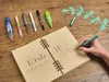 A hand is writing in a notebook with several pens and correction tape laid out on a wooden table. Anteprima dell’immagine prodotto