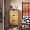 A narrow shoe cabinet with a geometric wood pattern and black frame, placed in a hallway next to a door and a window with blinds. A chair with a patterned cushion and a pair of boots are also visible. Produktbild-Vorschau