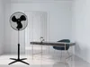 A black stand fan is shown in a modern, minimalist room with a desk and chair. Voir les images du produit