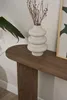 A white vase with dried branches sits on a wooden console table next to a large green leaf plant and a framed picture. Voir les images du produit