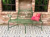 A green metal garden bench with a decorative pillow sits on a patterned tiled patio in front of a brick wall with two windows. Náhľad obrázku produktu