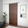 A modern sliding door with a wooden finish and silver hardware, mounted on a wall in a room with a plant and artwork. Produktbild-Vorschau