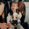 A young child is sitting in a car seat, smiling. There are numbered arrows overlaid on the image, highlighting features of the car seat. Produktbild-Vorschau
