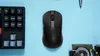 A black wireless mouse is centered on a light blue surface, with a portion of a black keyboard on the left and some small metallic objects on the right. Voir les images du produit
