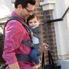 A man is carrying a baby in a carrier while walking outdoors near a brick wall and stairs. Anteprima dell’immagine prodotto