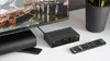 A black DVB-T2 tuner and remote control are placed on a white surface next to a television. Náhled obrázku produktu