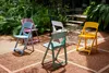 Several colorful modern folding chairs are arranged on a gravel patio with lush green foliage in the background. Produktbild-Vorschau