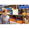 A person in a warehouse setting, with shelves of storage bins and boxes in the background. A display system with documents is mounted on a shelf. Náhled obrázku produktu