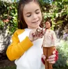 A young girl is holding a Barbie doll and a small monkey toy outdoors. Produktbild-Vorschau