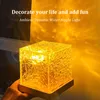 A cube-shaped lamp with a textured surface, emitting a warm yellow light that casts ripple patterns on the background. A hand is holding the lamp from the bottom. Produktbild-Vorschau