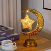 A decorative table lamp shaped like a crescent moon with a star hanging from it, placed on a wooden table next to books and a cup. Produktbild-Vorschau