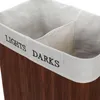 Close-up view of the top opening of a two-compartment laundry basket made of dark wood slats with a white fabric lining. The words "LIGHTS" and "DARKS" are embroidered on the fabric rim. Produktbild-Vorschau