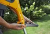 A person's hands are shown adjusting a yellow garden trimmer in an outdoor setting with green grass and bushes in the background. Produktbild-Vorschau