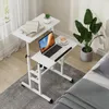 A mobile laptop table with a laptop, coffee cup, and books is placed next to a bed, with a large plant in the background. Podgląd zdjęcia produktu