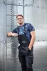 A man wearing black overalls and a blue camouflage t-shirt leans against a large metal silo with a ladder. Náhled obrázku produktu