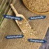 A wooden scoop filled with small grains, placed on a burlap cloth, with text overlays indicating product features. Produktbild-Vorschau